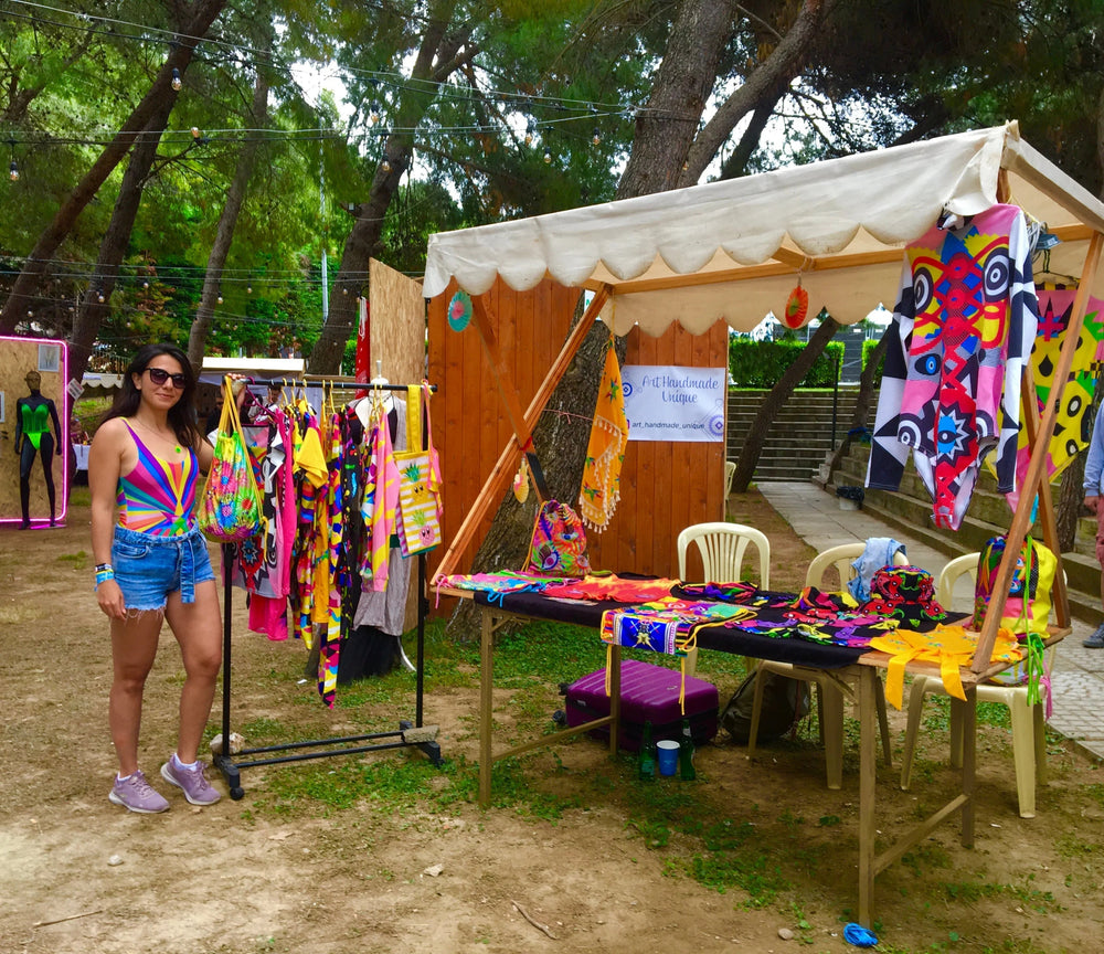 Woman standing next to colorful handmade clothing and accessories at outdoor market stall