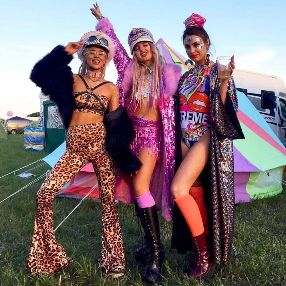 Three women in colorful rave outfits posing outdoors at a festival with tents and camper in background