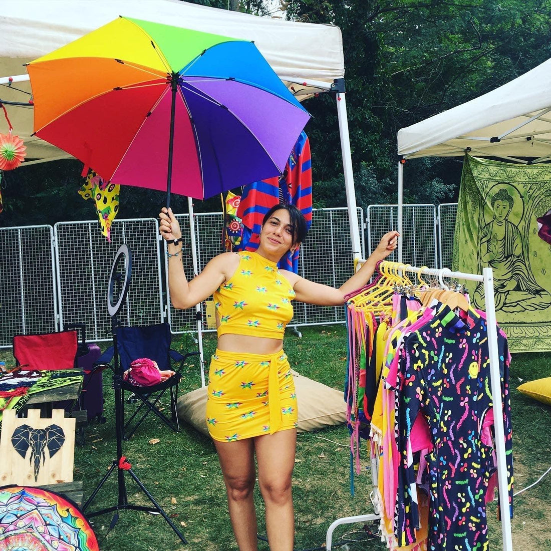 Woman in yellow star print outfit holding rainbow umbrella at outdoor market stall with colorful clothes
