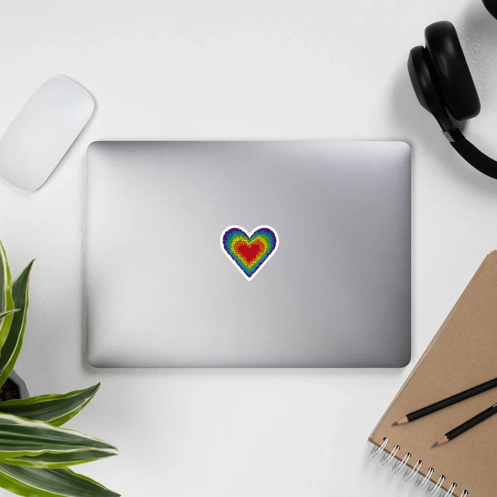 Silver laptop on white desk with colorful rainbow heart sticker, wireless mouse, black headphones, plant, and notebook