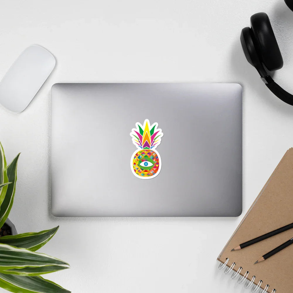 Silver laptop with colorful pineapple sticker featuring an eye, on white desk with mouse, headphones, notebook, pencils, and plant
