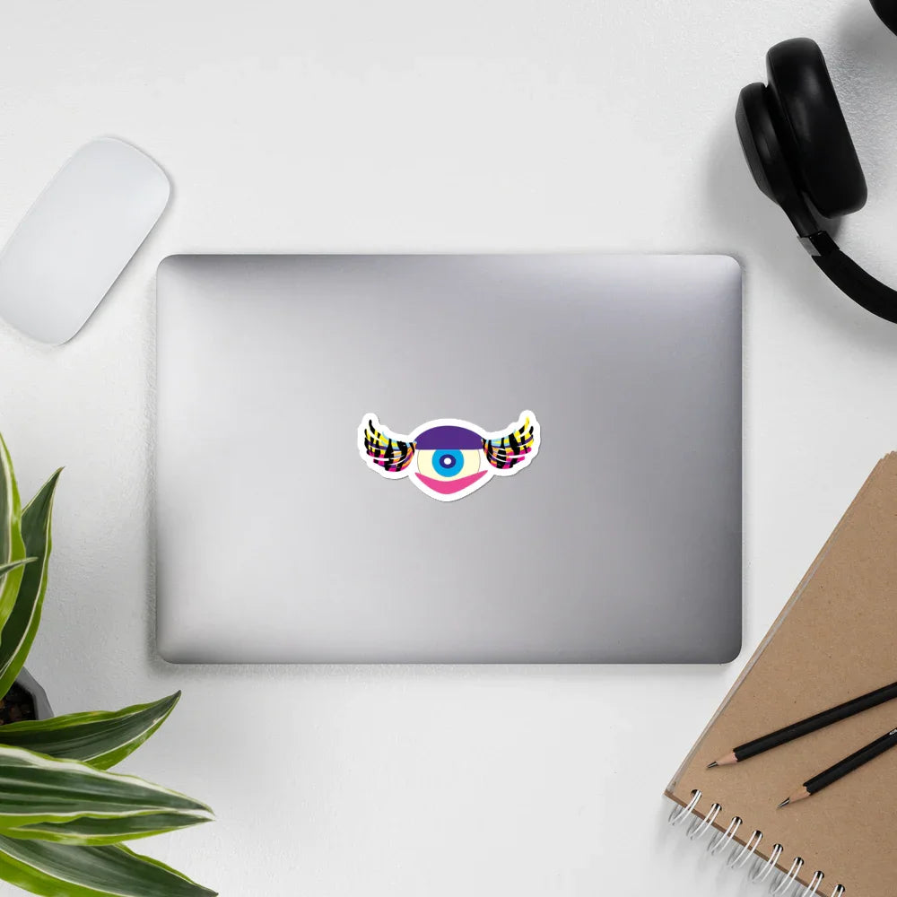 Silver laptop with colorful eye sticker with wings on white desk, surrounded by headphones, mouse, notebook, and plant